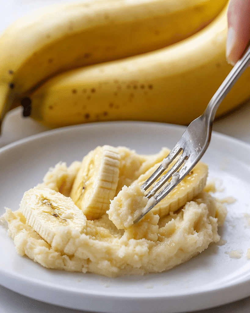 Gluten Free Banana Nut Bread Muffins: A Complete Guide 8 Mashed bananas on a white plate with a fork, used as an ingredient for gluten free banana nut bread muffins, with ripe bananas in the background.